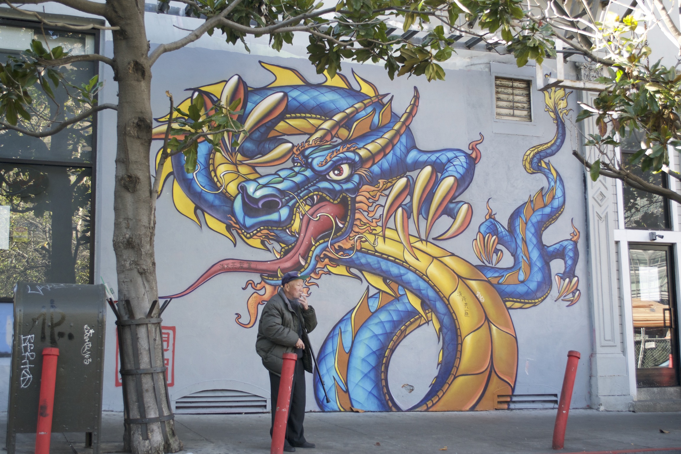An old smoking man sands in front of a mural of a blue dragon.
