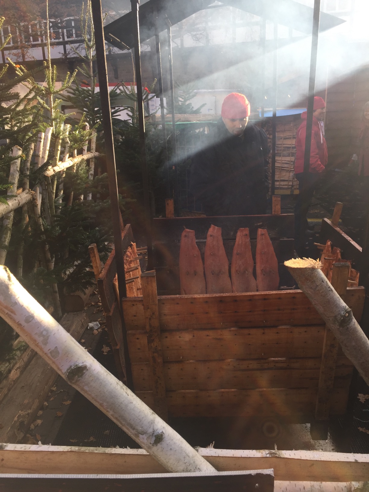A man looks at a smoking pit surrounded by planks with strips of fish hanging from them.