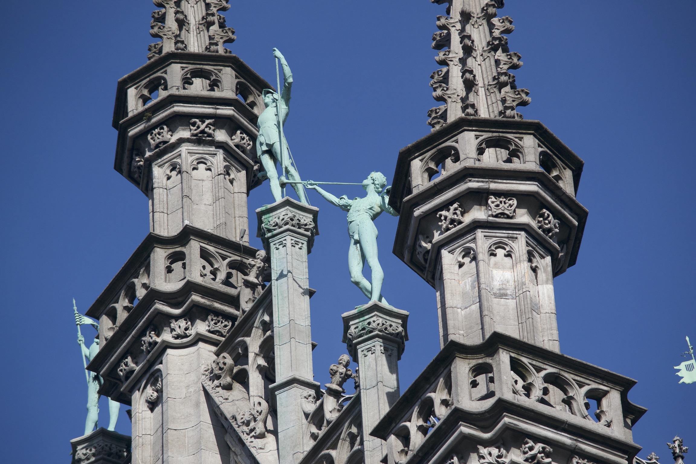 Two copper-green statues stand atop ornate pedestals amidst two spires.  One holds a pike, the other blows a herald's trumpet.