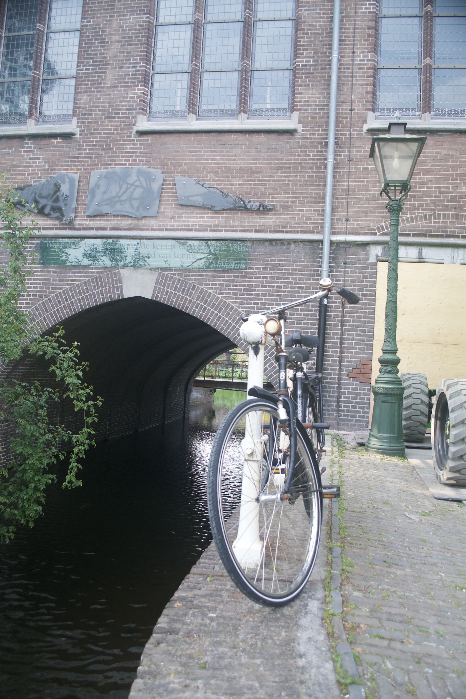 A bicycle locked to a post on the edge of a canal which goes through a tunnel in a building in the background.