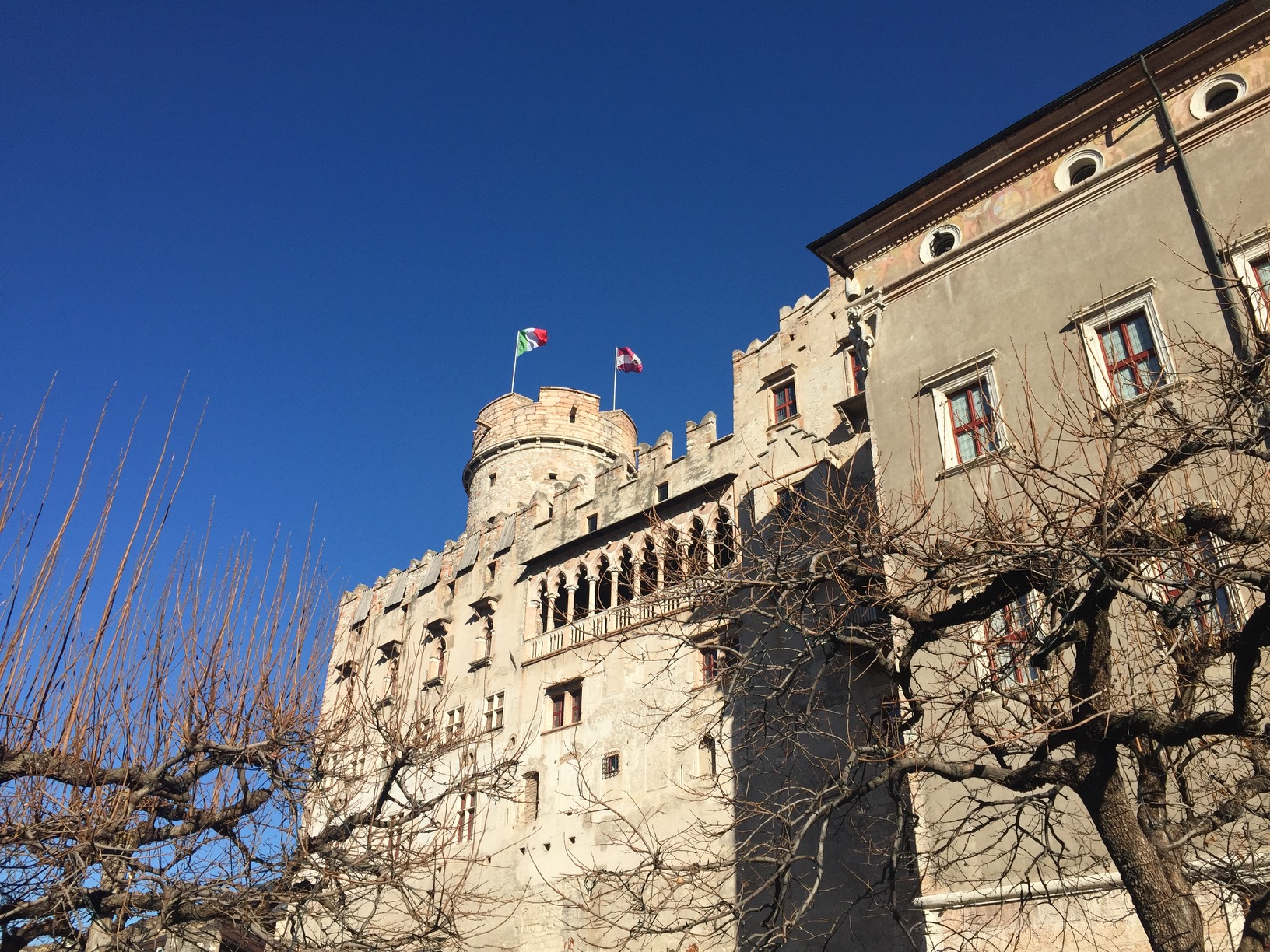A large, rectangular building in two architectural styles, with a round tower flying the Italian tricolor and the red-and-white flag of Trento province.