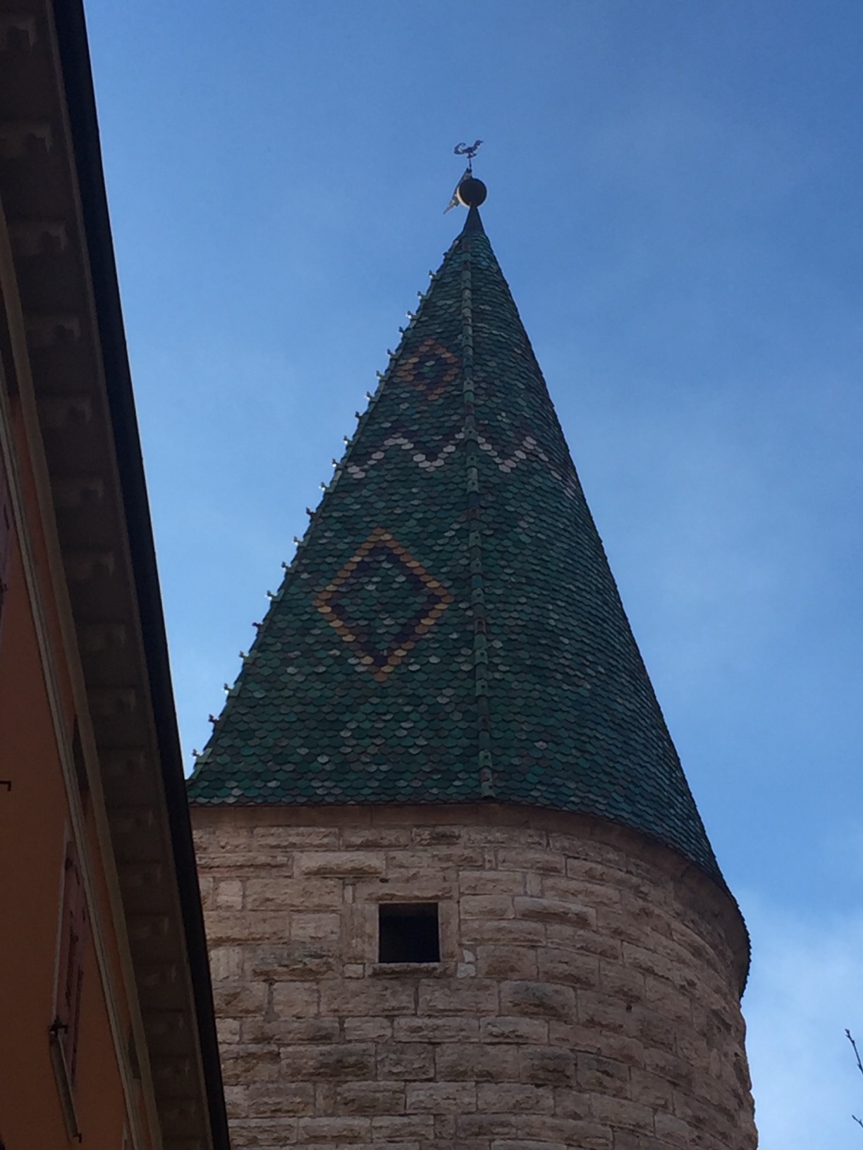 The top of an old tower of brown stones, with a steep, patterened green tile roof.