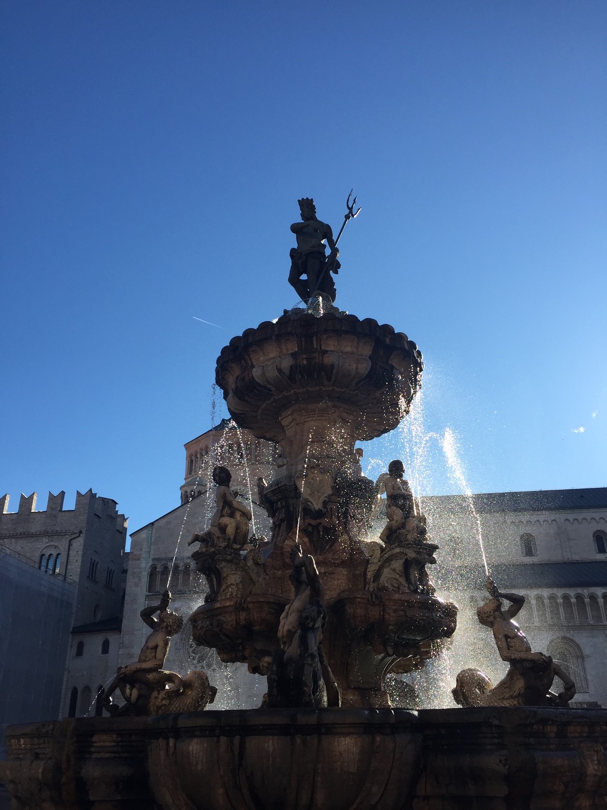 A stone fountain of mermaids spewing water towards a bronze trident-wielding Neptune.