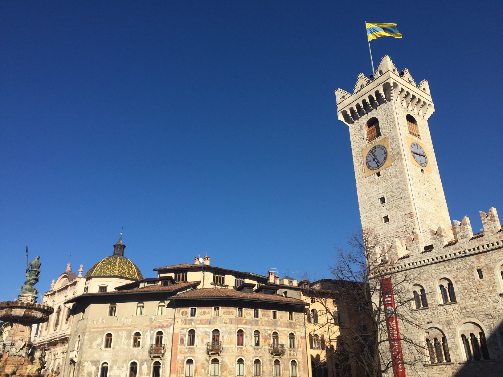 A square clock tower flying the yellow-and-blue flag of of Trento city, with a round tiled tower and fountain of Neptune nearby.