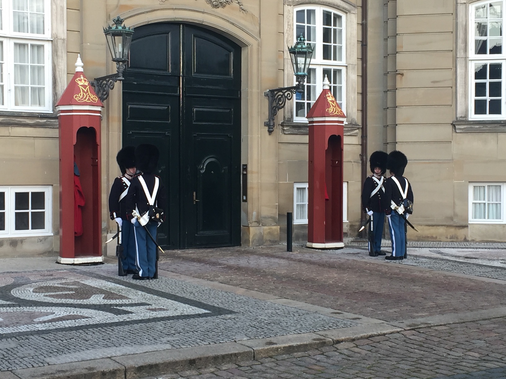 Two pairs of guards dressed in blue pants, black jackets, and tall, furry hats, face one another before two red booths a wide, black doorway into a stone building.