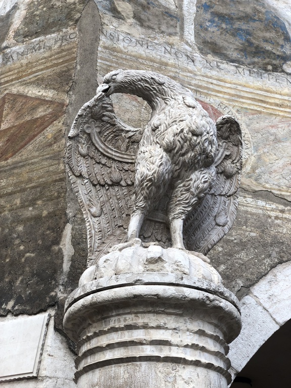 A stone eagle on a pedastal, preening its wings, in front of a dirty fresco.