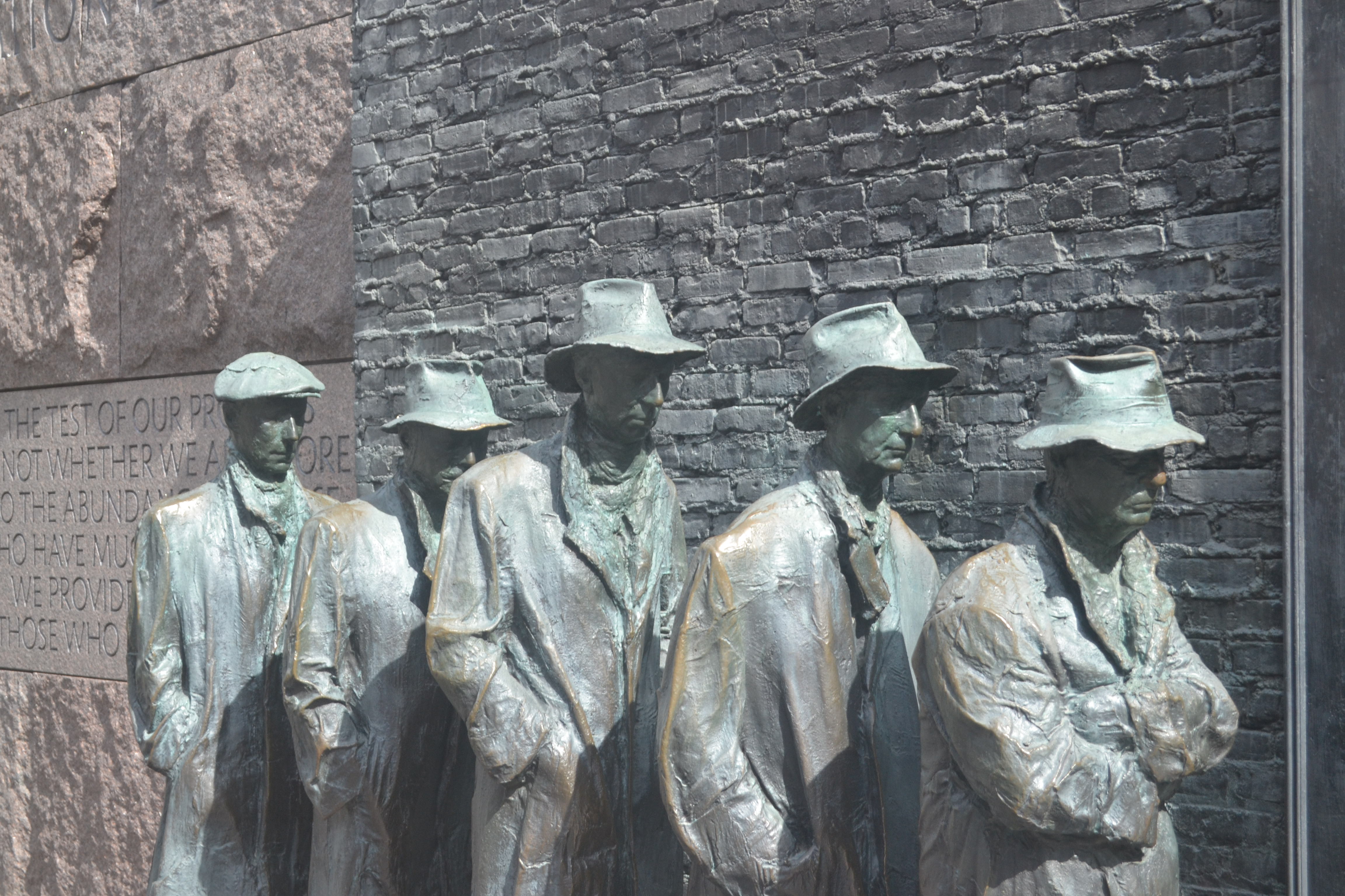 Statues of depressed bronze men stand single-file before a gray brick wall.