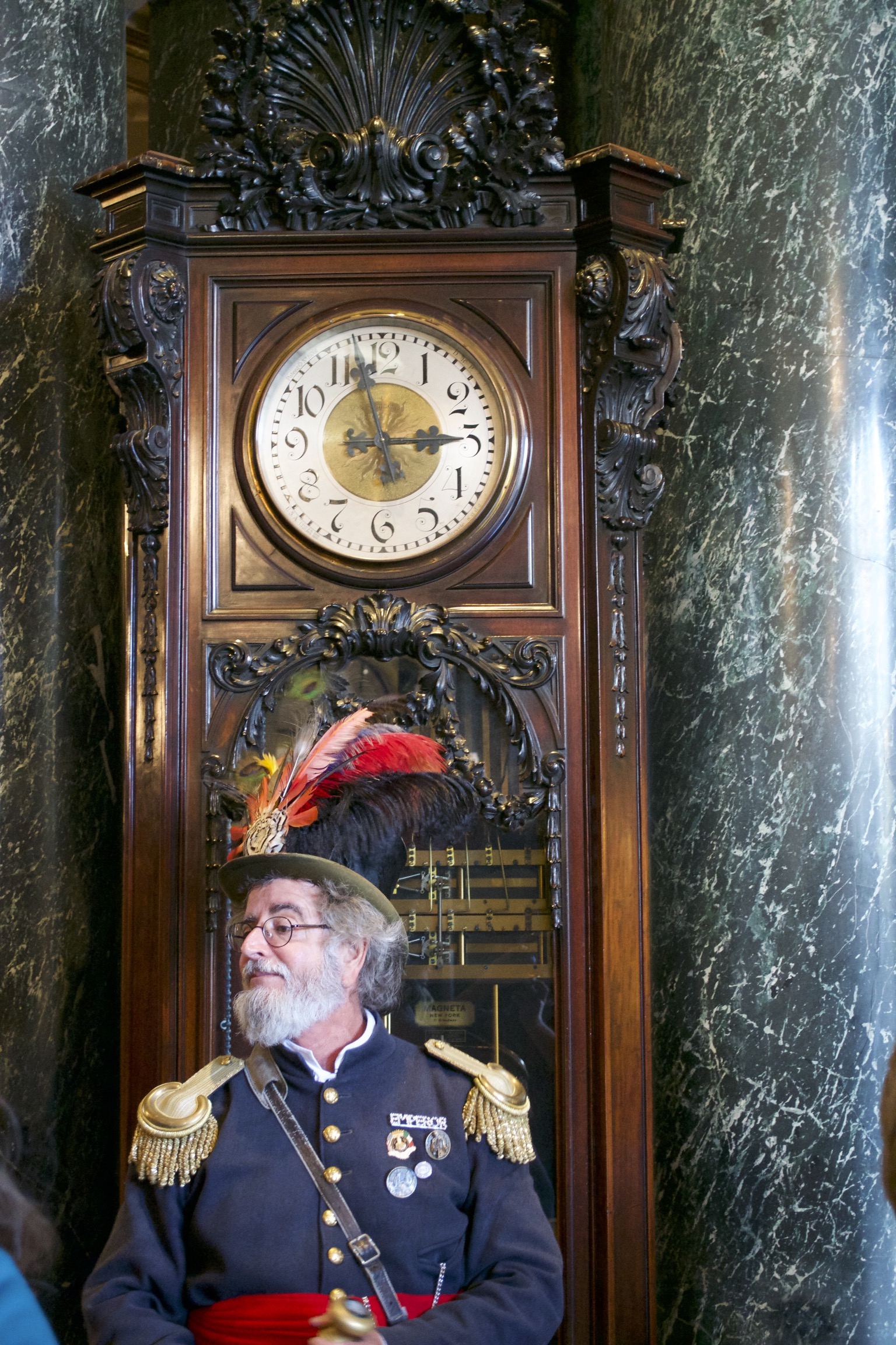 A man in a navy blue officer's jacket with gold epaulets, a red cumberbund, and a feathered top hat stands in front of a huge grandfather clock.