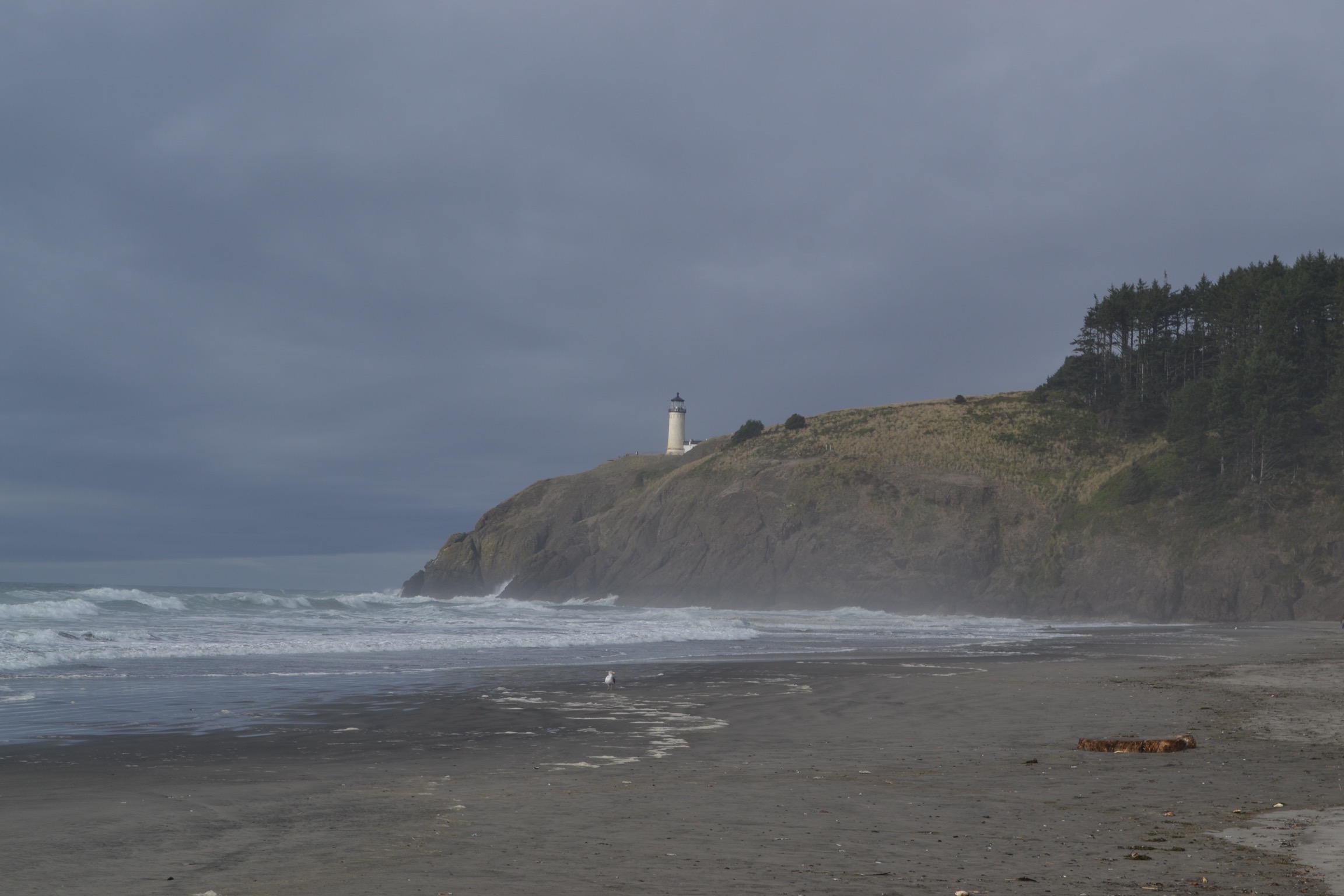 A beach with rough waves and cliffs with a plain white lighthouse before a gray sky.