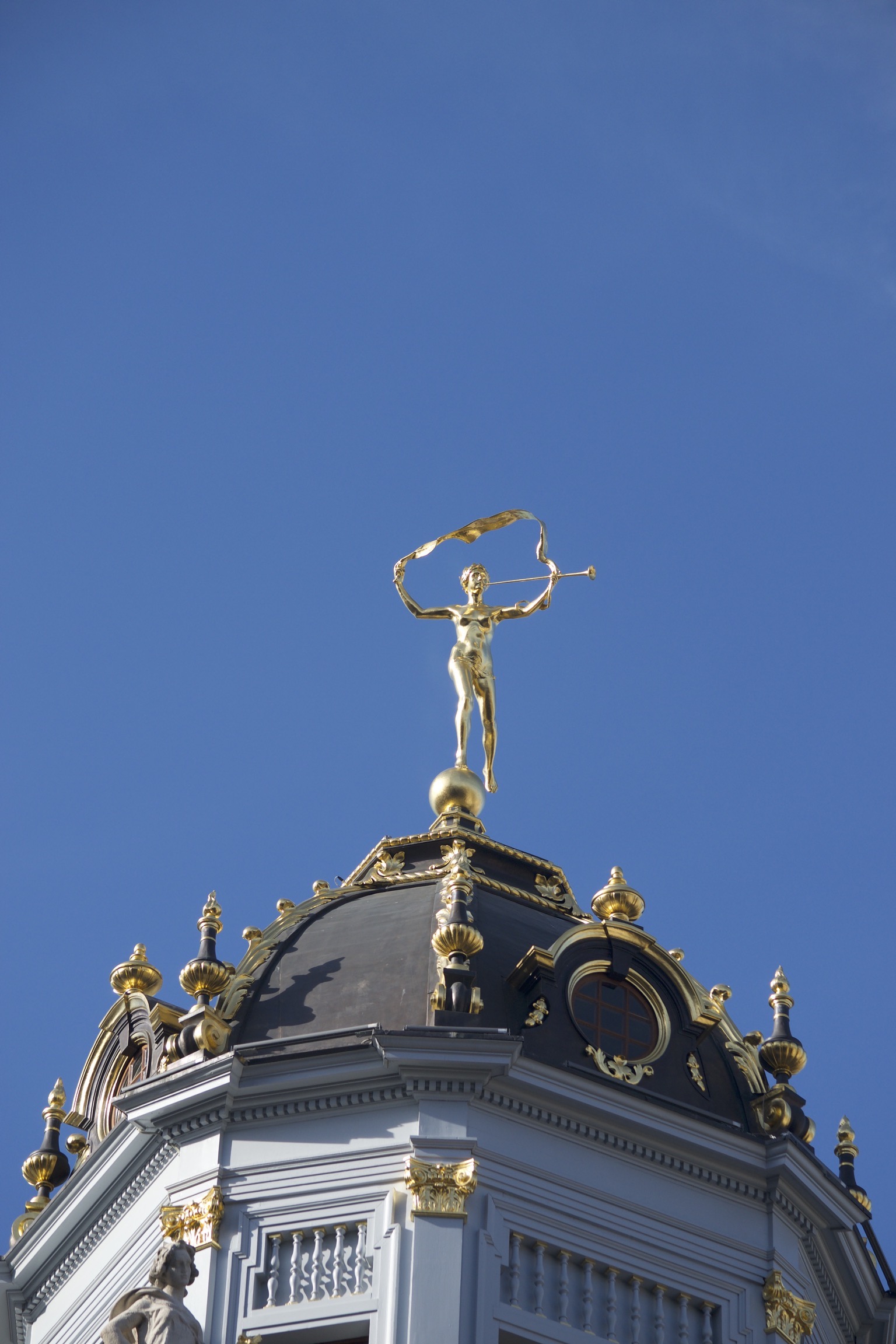 A golden figure blows a trumpet and holds a long ribbon atop a black and gilted roof dome.
