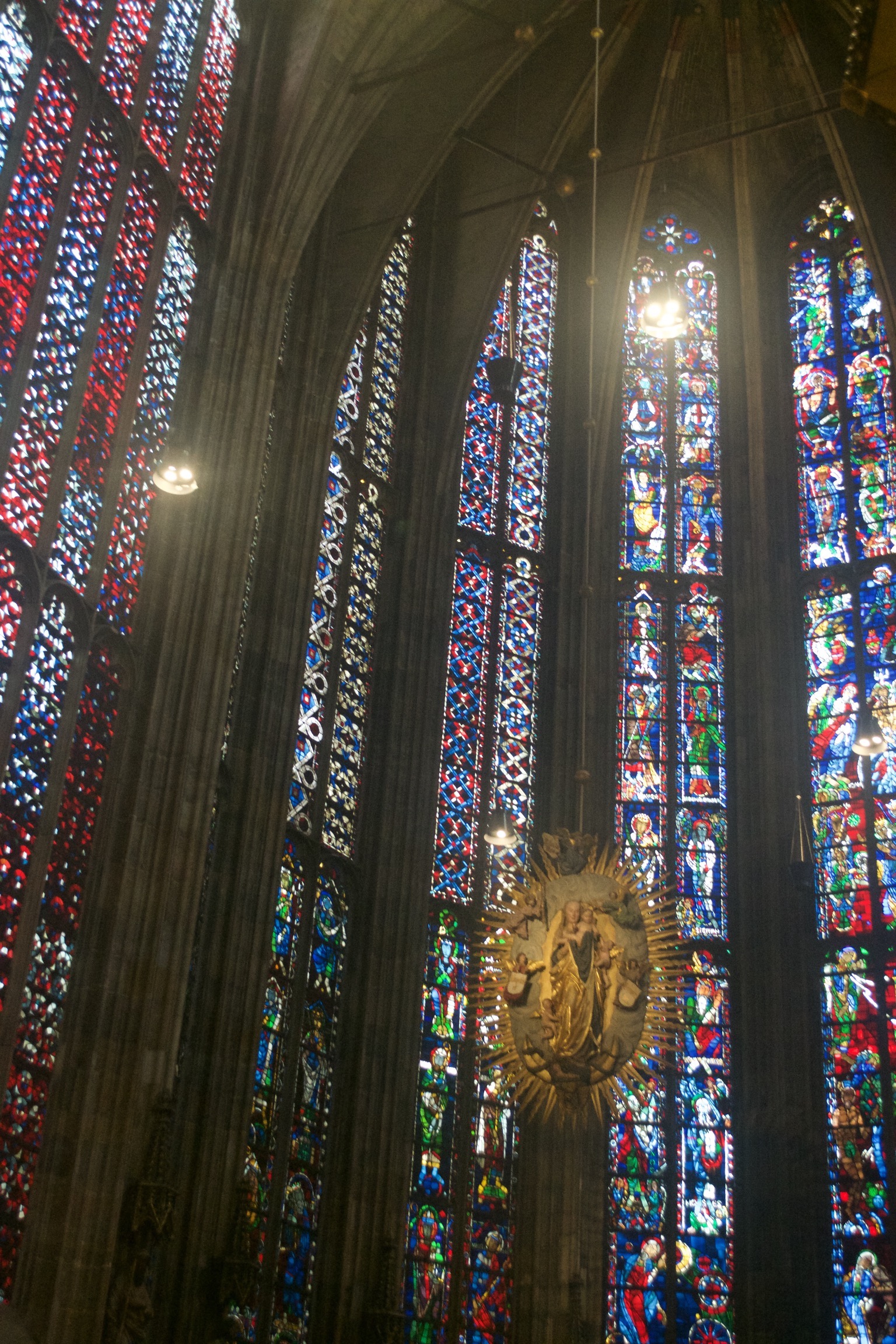 The apse of a Gothic cathedral illuminated with stained glass.  An emblem of Mary and the infant Jesus hangs from the ceiling.