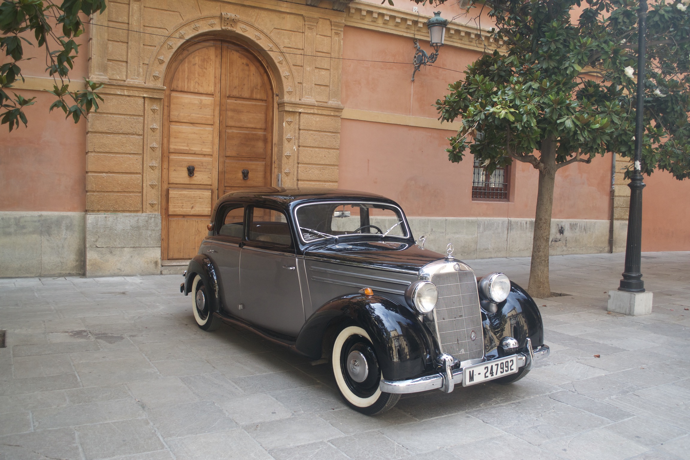 A classic black and gray Mercedes sits in a plaza before a Spanish building's large wooden doors.