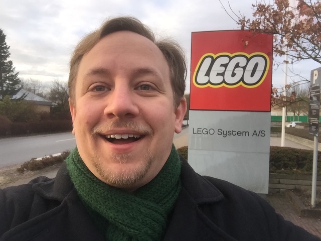 A 30-year-old man with blond hair and beard, smiling widely in front of the red LEGO sign of the LEGO Headquarters.