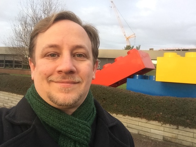 The same goofy man in front of a statue of three giant, adult-sized, four-by-two LEGO bricks.
