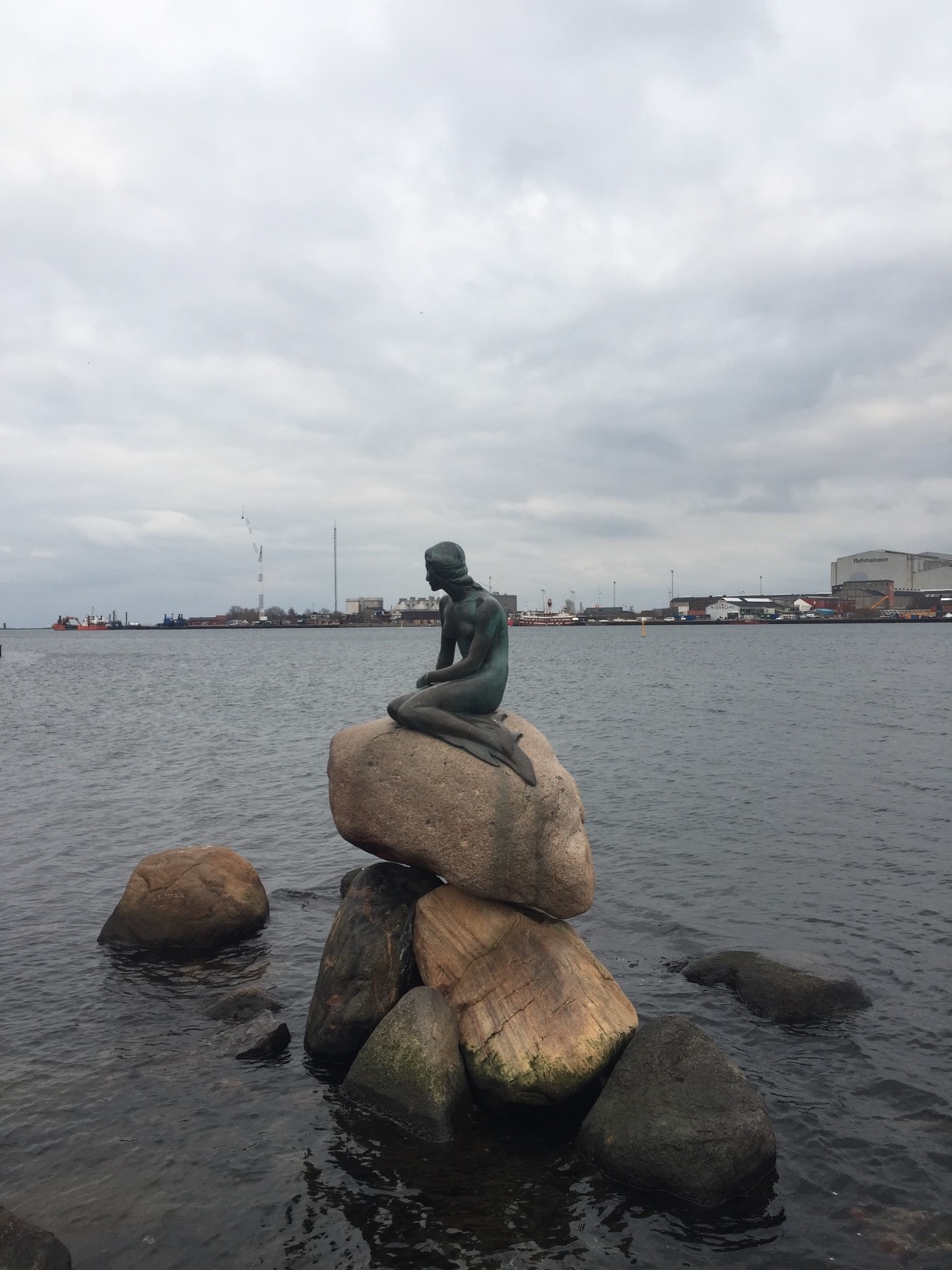 A bronze statue of a female mermaid rests on a rock in a harbor.