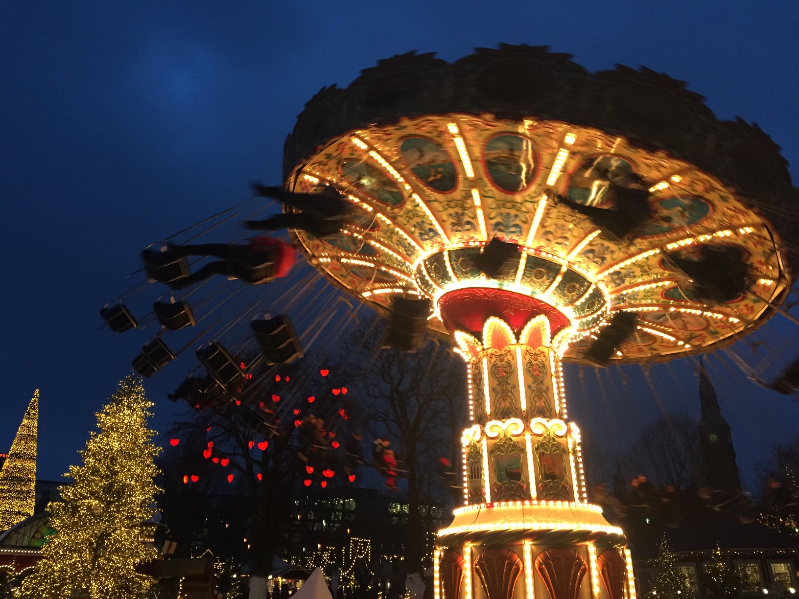 A brightly-lit swing carousel near an illuminated Christmas tree before a dark sky.