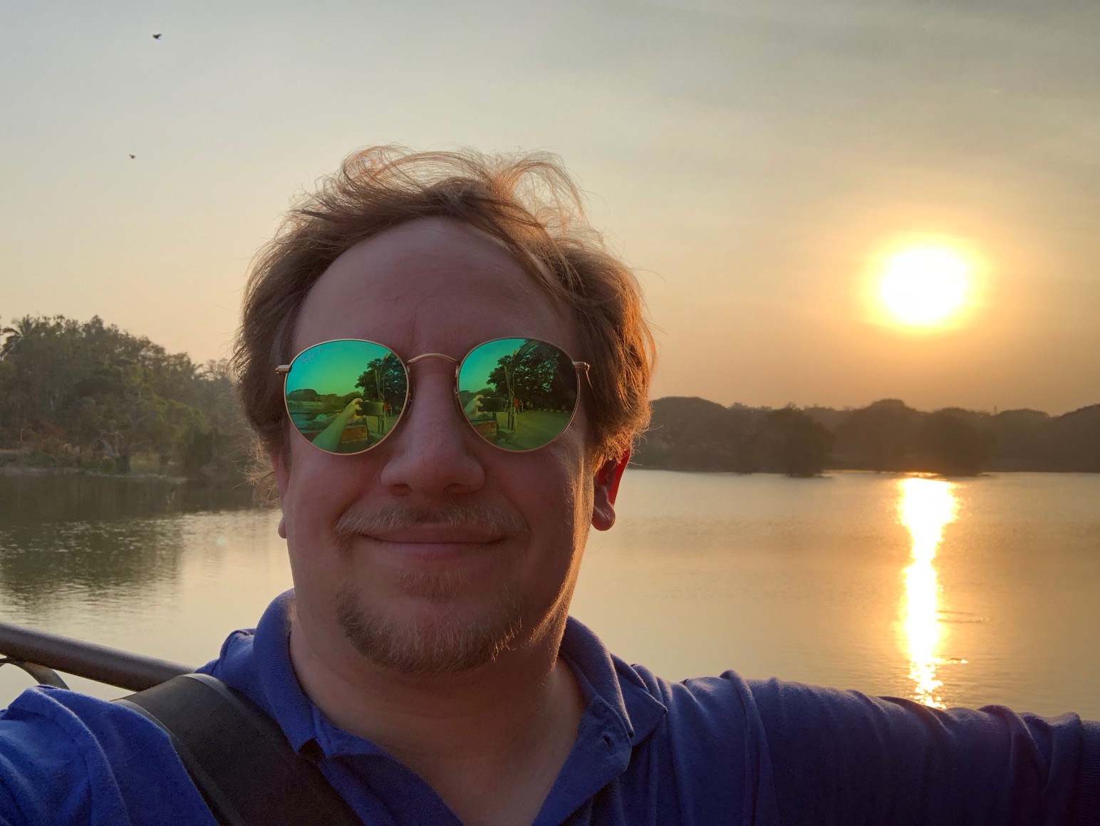 The sun sets over a lake, behind a man with a goatee, messy blond hair, and preposterous sunglasses.