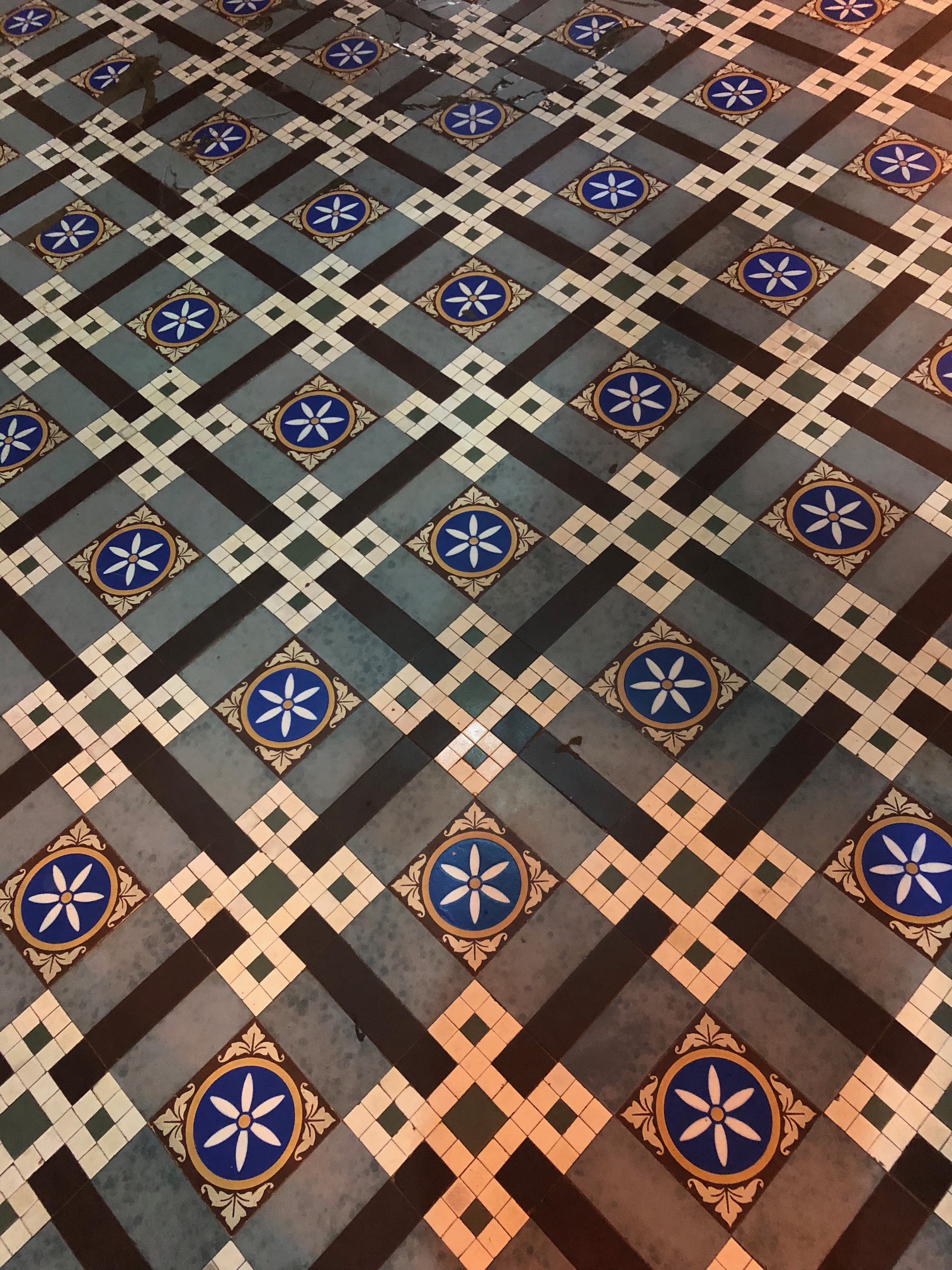 An intricate square pattern of floor tiles, including white six-petaled flowers on a blue background and small off-white tiles arranged in a heraldic knot.