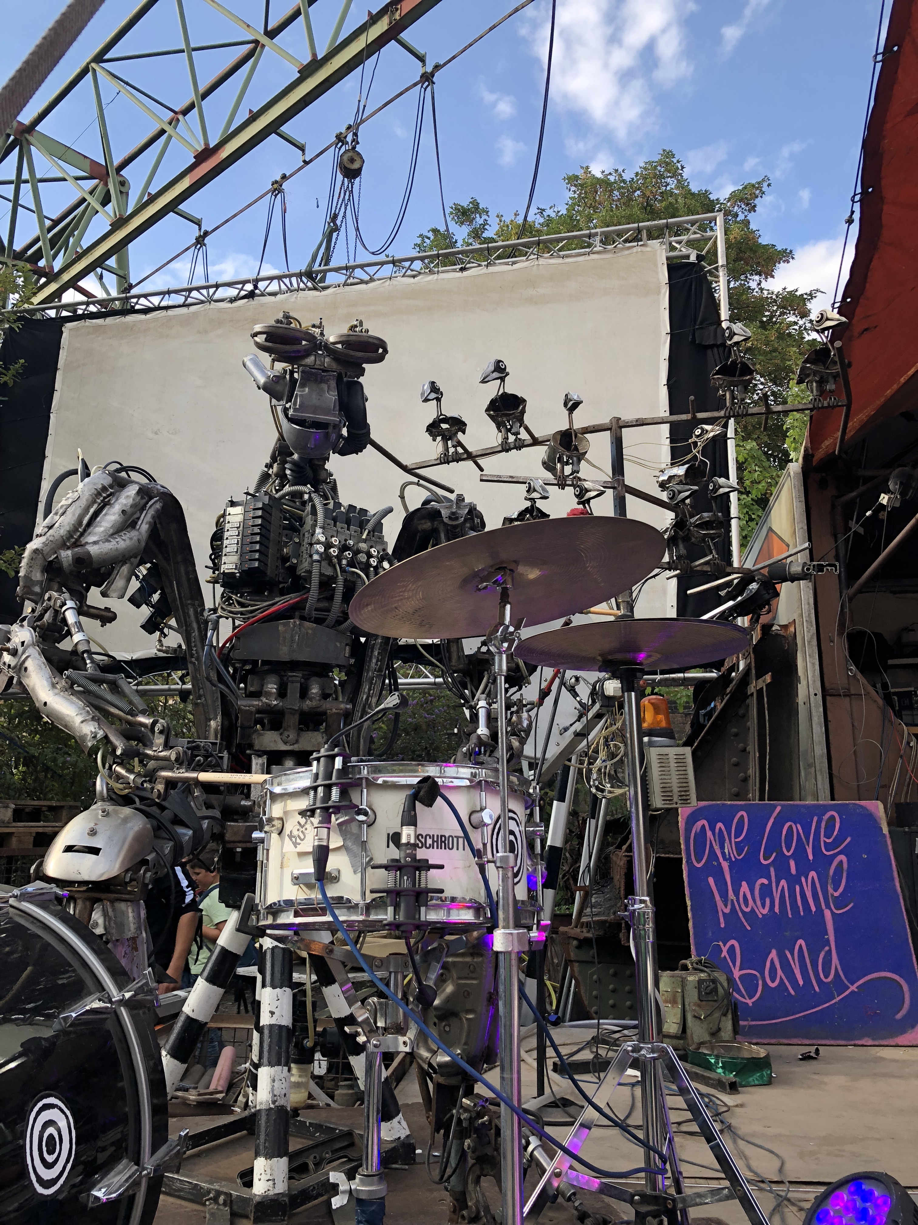A robot sitting behind a drum set, with a sign that reads 'One Love Machine Band'