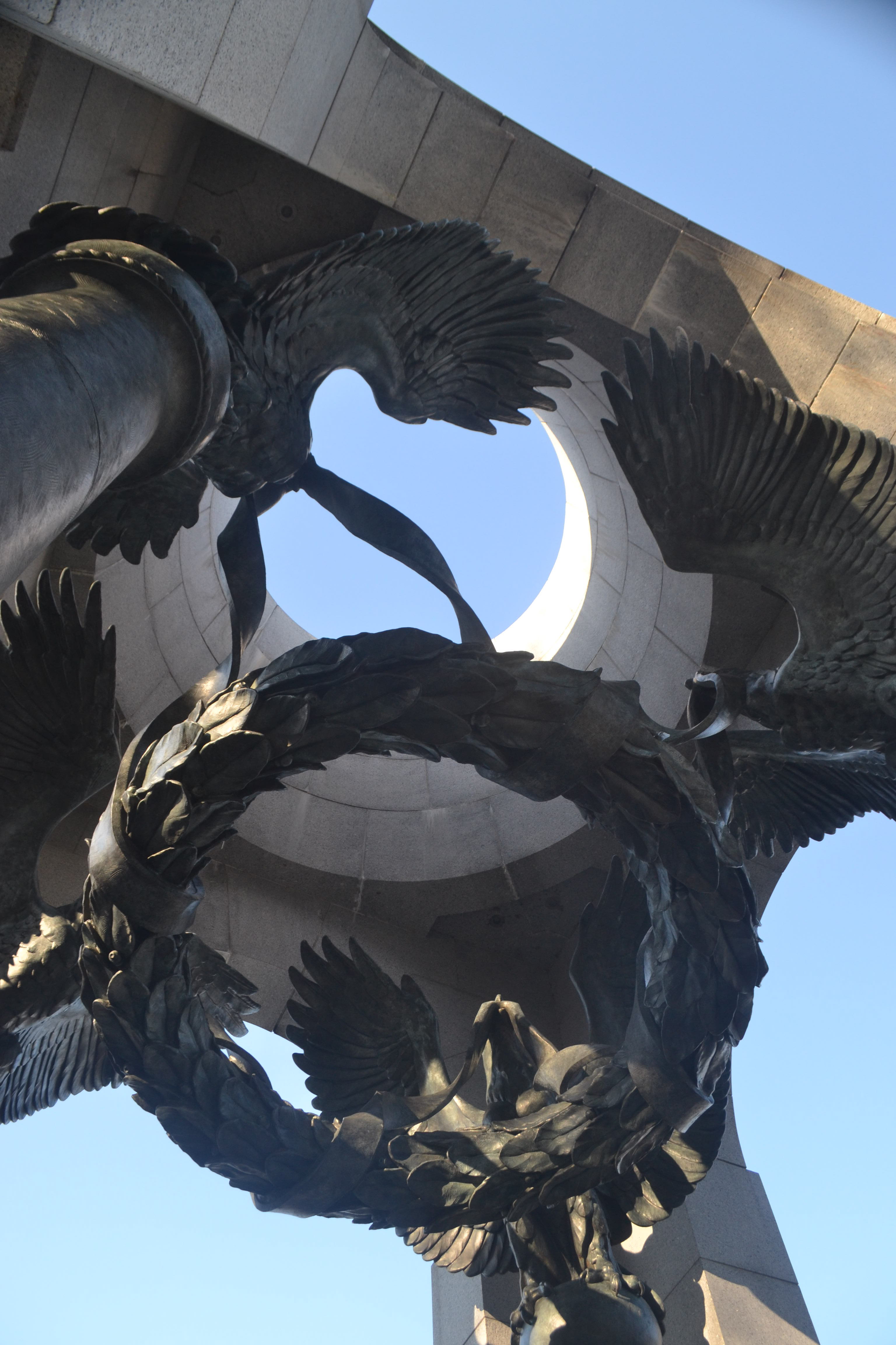 Four metallic eagles, wings spread, support a wreath by a ribbon in their beaks, beneath a stone, triumphal arch.