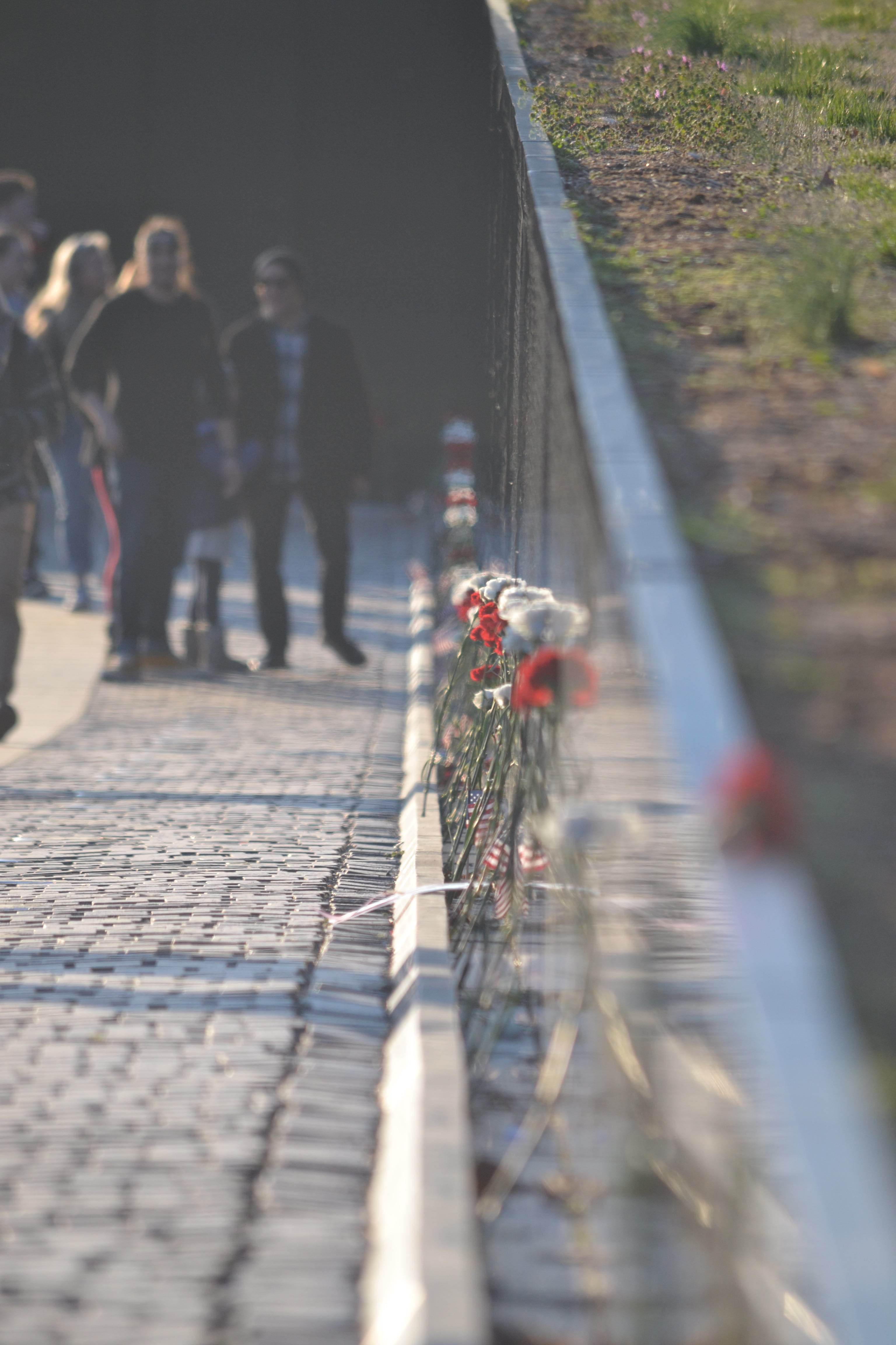 A stone walkway descends parallel to a black mirrored wall with red and white flowers and small flags leant against it.